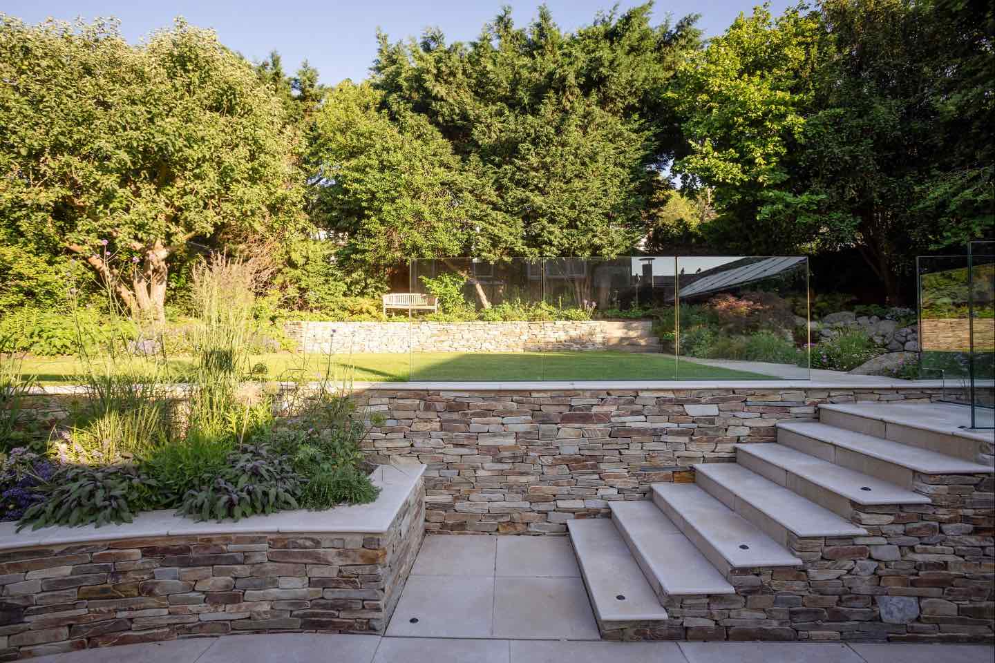Country walled garden in Hertfordshire with retaining wall clad with yellow quartz paddle stones and frameless glass balustrade for safety