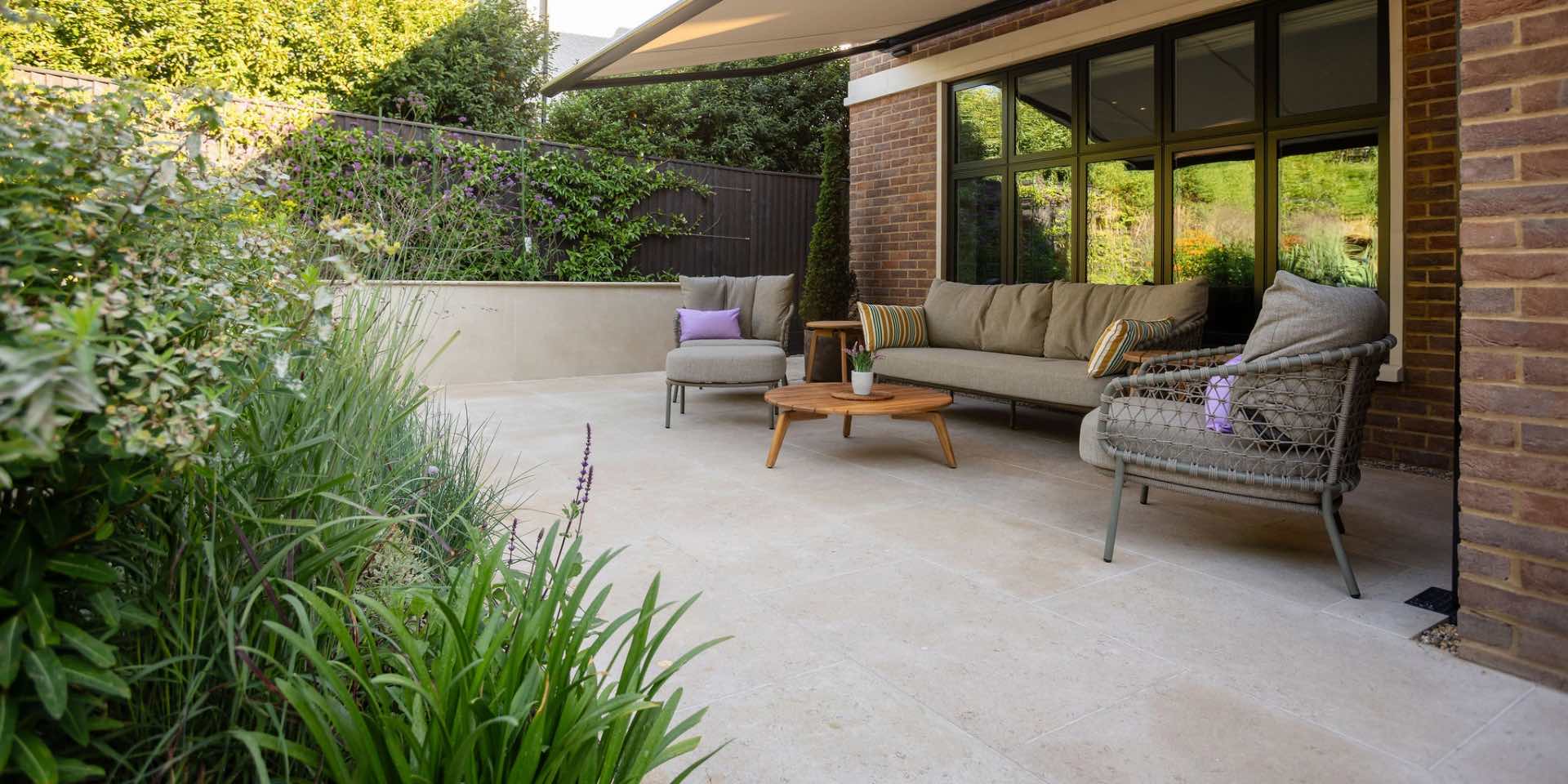 Relaxed outdoor lounge set with sofa and armchairs in richly planted garden with limestone paving and overhead awning for shade