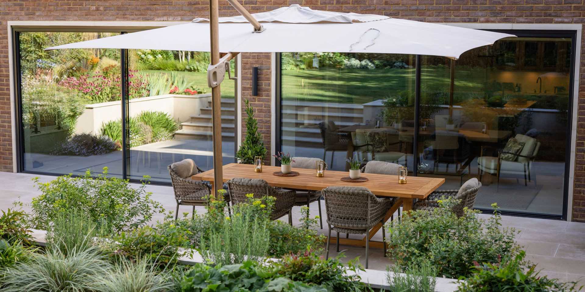 Large cantilever parasol with large teak dining table and chairs, on sunken limestone patio surrounded by lush, colourful planting for sun