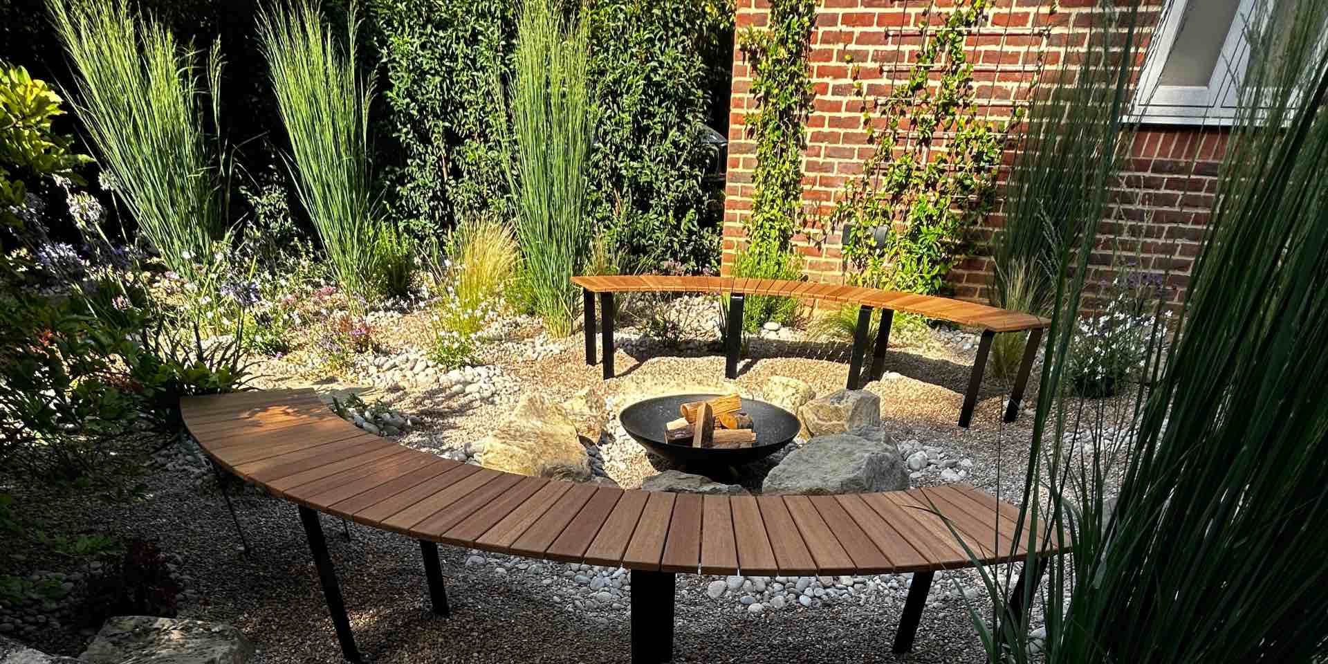 Timber firepit seating with curved seats around a circular cast iron fire pit set within an area of boulders and pebbles with sunny mediterranean planting