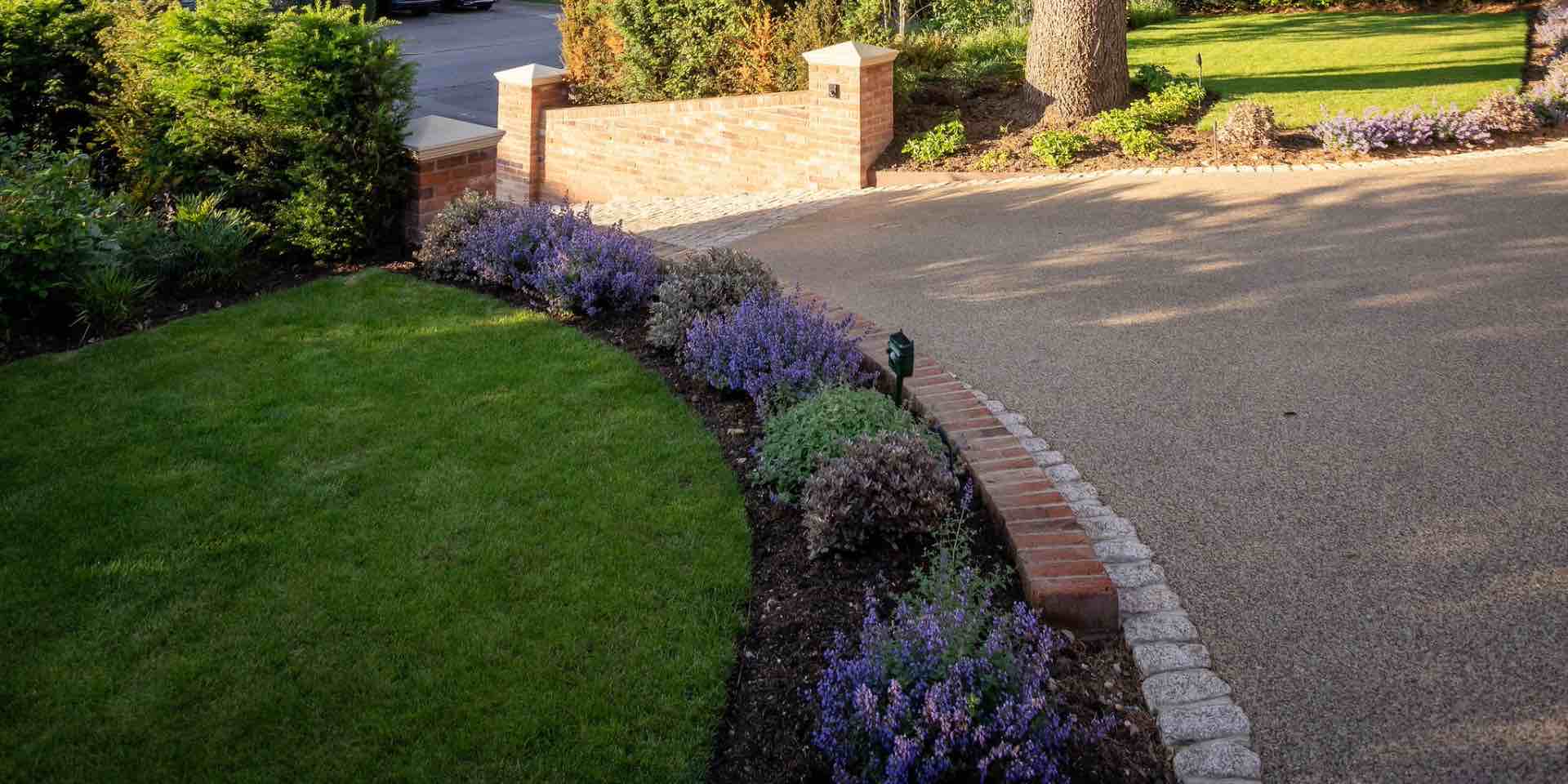 Resin bound drive with steep entrance and yellow tumbled granite sett highway crossover with red brick retaining walls and pier caps with adjoining planting and lawn